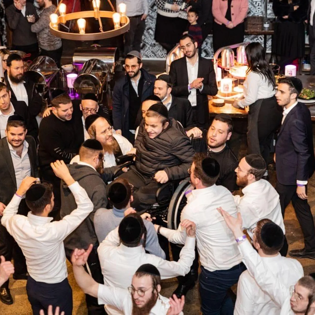 Group of men, many wearing traditional Jewish head coverings, celebrating at an indoor event with one person in a wheelchair in the center being lifted up and cheered around.
