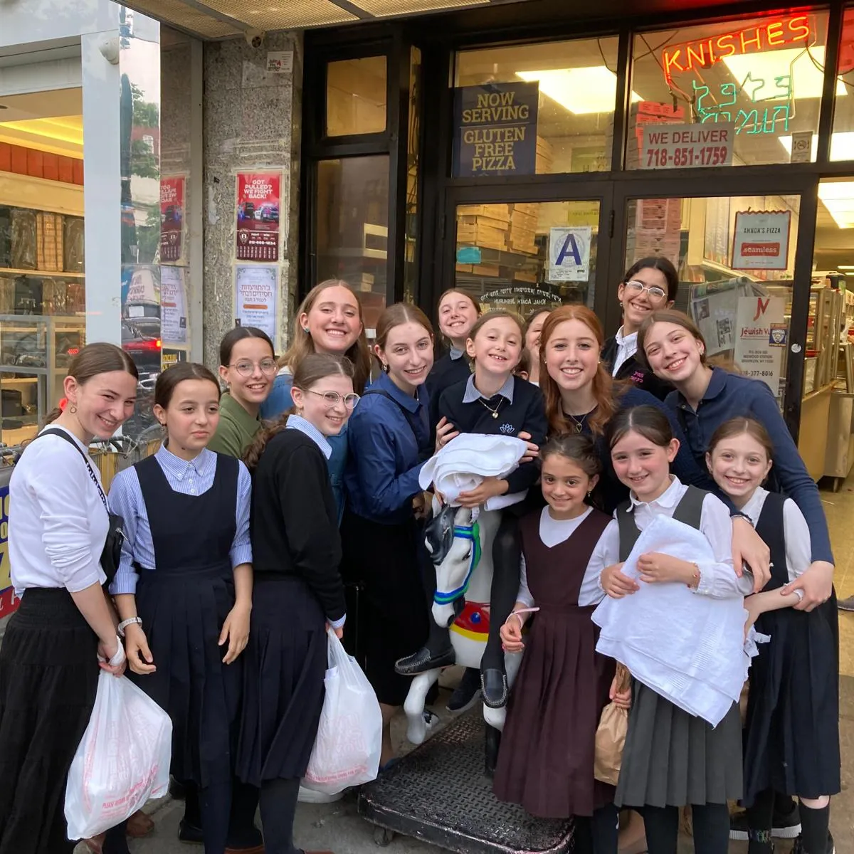 Group of smiling girls and young women standing in front of a storefront with signs advertising gluten free pizza, knishes, and delivery, some holding shopping bags and a few gathered around a small coin-operated horse ride.