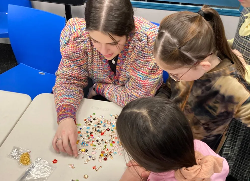 Three girls sitting around a table sorting or playing with a variety of small colorful charms and beads scattered on the table surface in a classroom setting.