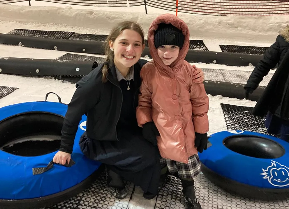 Two girls dressed in winter clothing pose for a photo outdoors on snow-covered ground, with large blue snow tubes nearby.