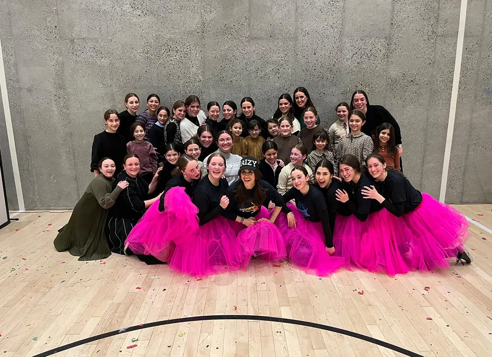 Group of 32 smiling girls and young women posing indoors on a wooden floor in front of a concrete wall, some wearing bright pink tulle skirts and others in dark or neutral clothing.