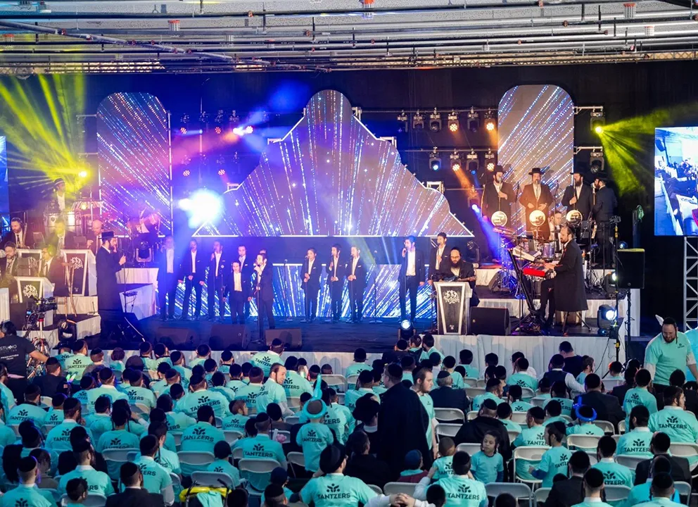 Large indoor concert with a stage featuring a vocal group and musicians performing in front of colorful LED backdrop panels, and a seated audience wearing matching turquoise shirts labeled 'Tantzers'.