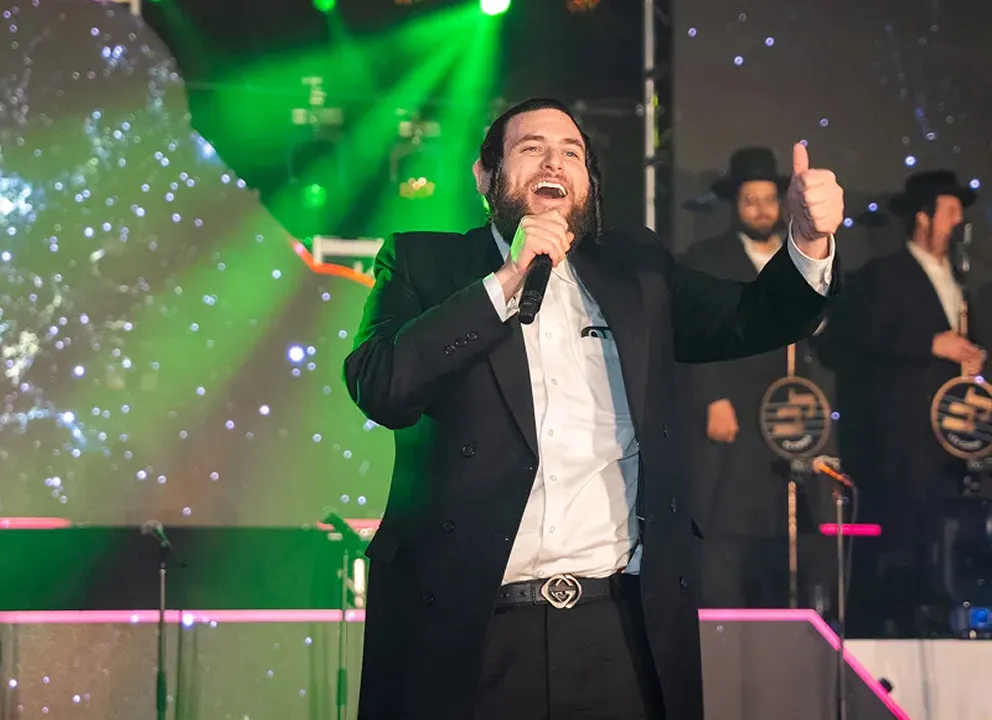 Man in dark suit and white shirt singing into a microphone on stage, giving a thumbs-up, with green stage lights and musicians in the background.