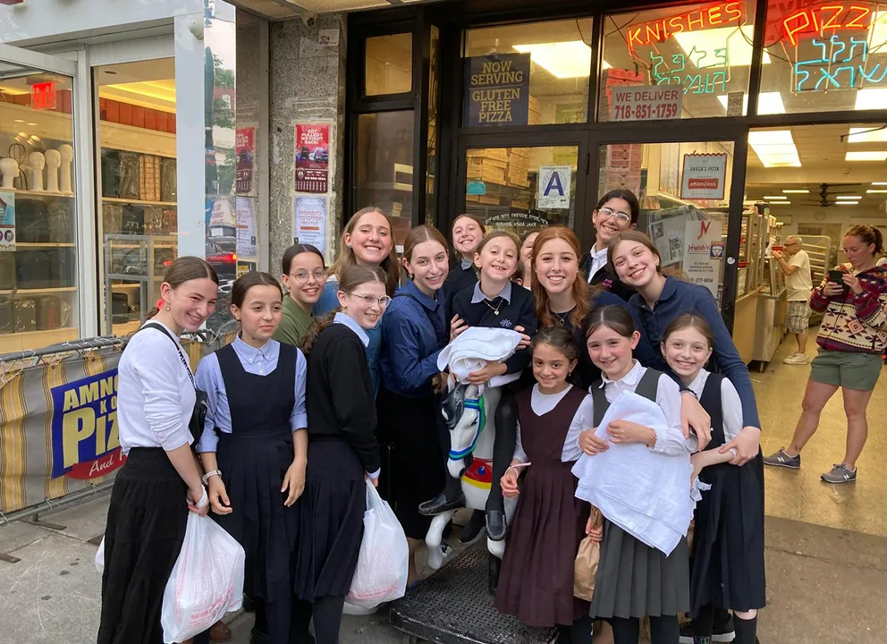 Group of smiling girls in modest school uniforms posing outside a pizza shop holding bags and towels with neon signs reading 'Knishes' and 'Pizza' visible inside.