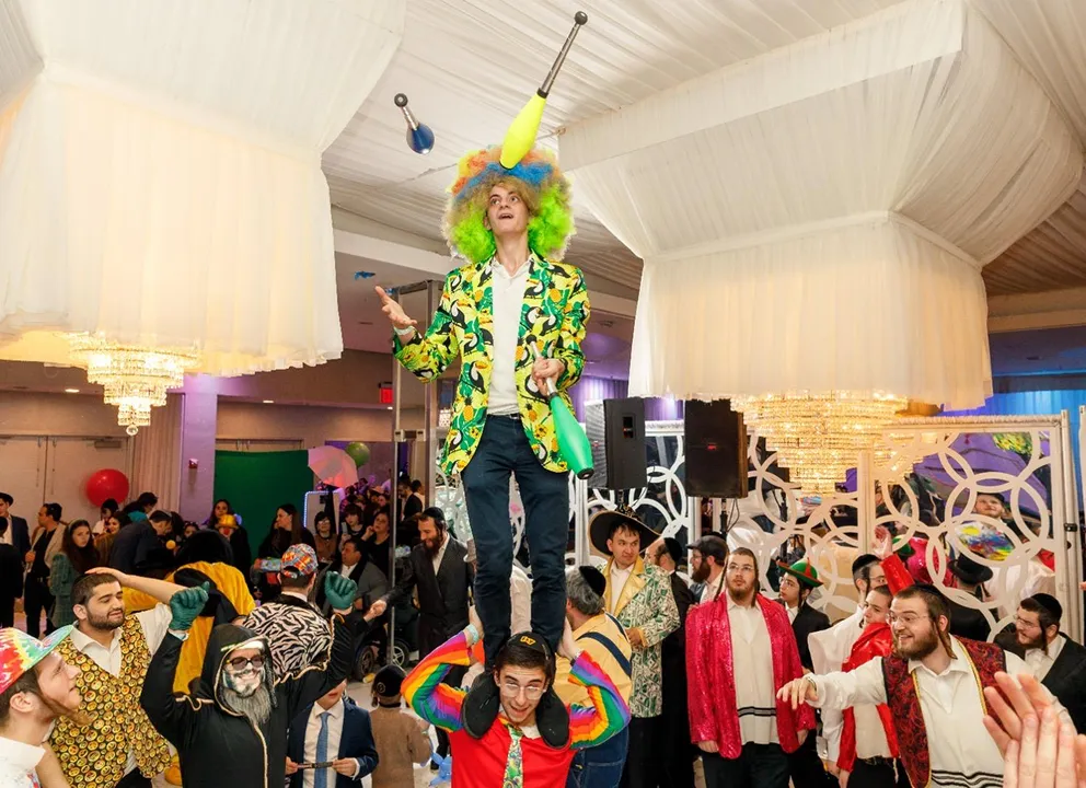 A lively indoor party scene with a man wearing a colorful clown wig and patterned jacket juggling clubs while standing on the shoulders of another person in a rainbow-striped sweater, surrounded by a crowd of people dressed in festive attire under large decorative chandeliers.