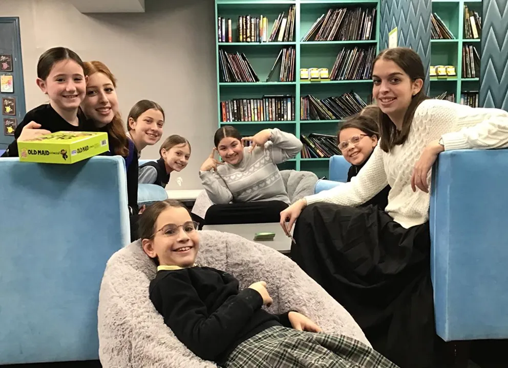 Group of eight smiling children sitting and lounging in a cozy room with blue furniture and bookshelves filled with books.