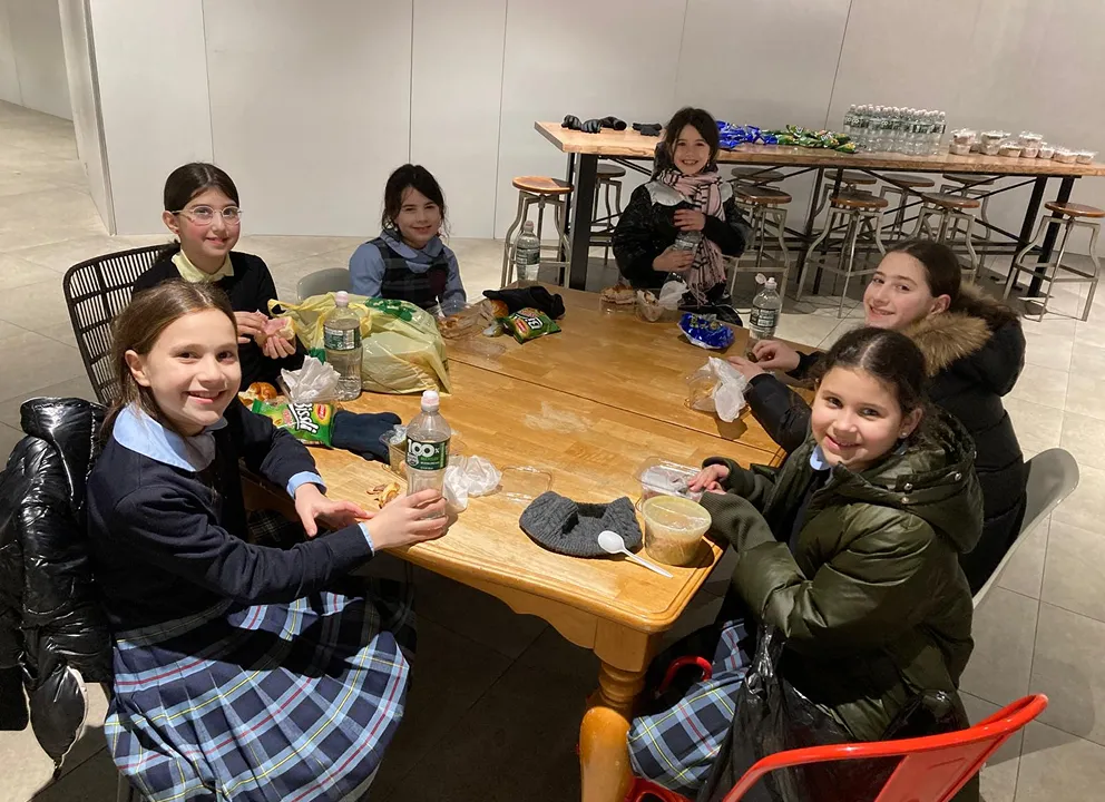 Six schoolgirls sitting around a wooden table indoors, eating snacks and drinking water, wearing plaid skirts and jackets, with food and water bottles on the table.