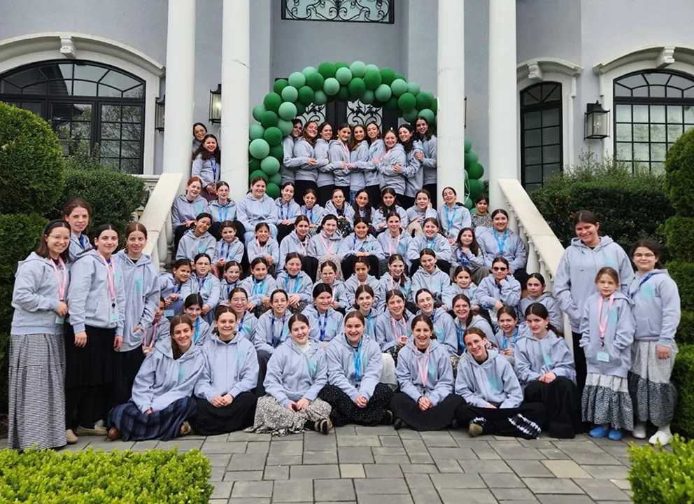 Large group of girls and young women wearing matching light gray hoodies posing on the steps of a building entrance decorated with a green balloon arch, surrounded by shrubs and windows.