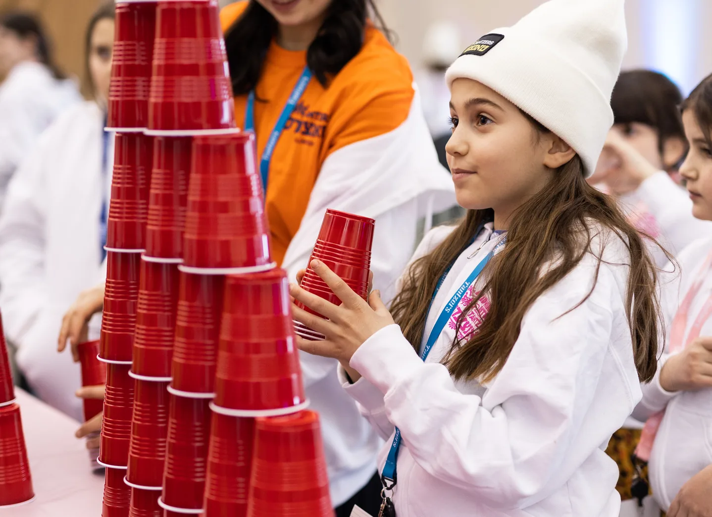 Girl in white sweatshirt and beanie stacking red plastic cups at an event with others nearby.