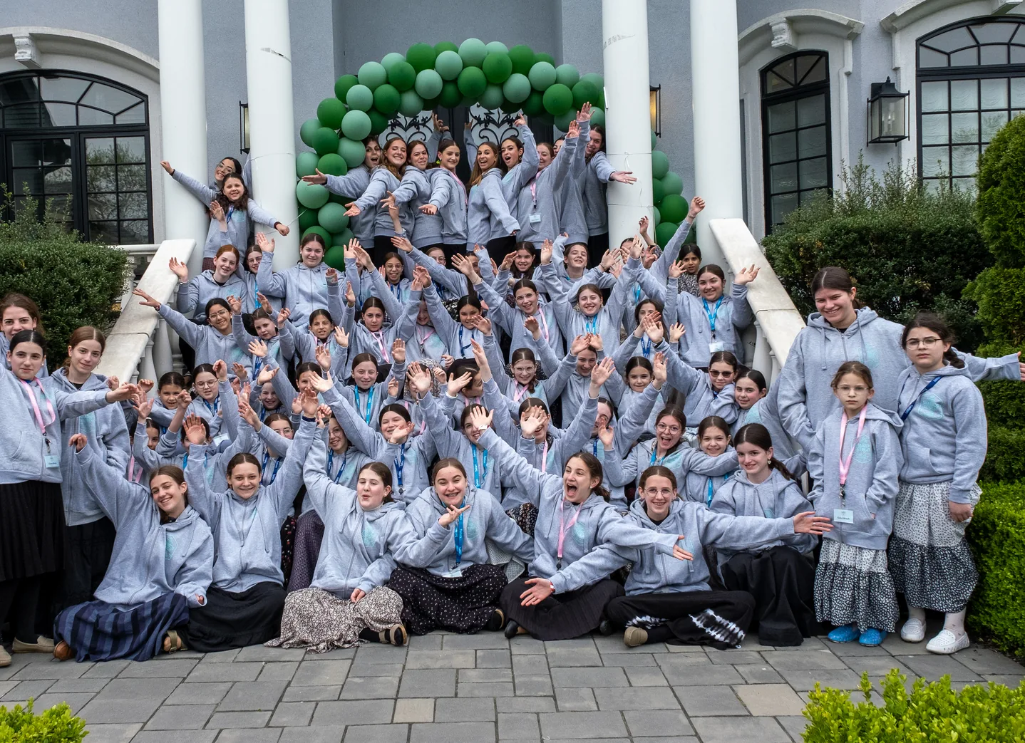 Large group of smiling girls in gray hoodies posing with raised arms outside building with green balloon arch.