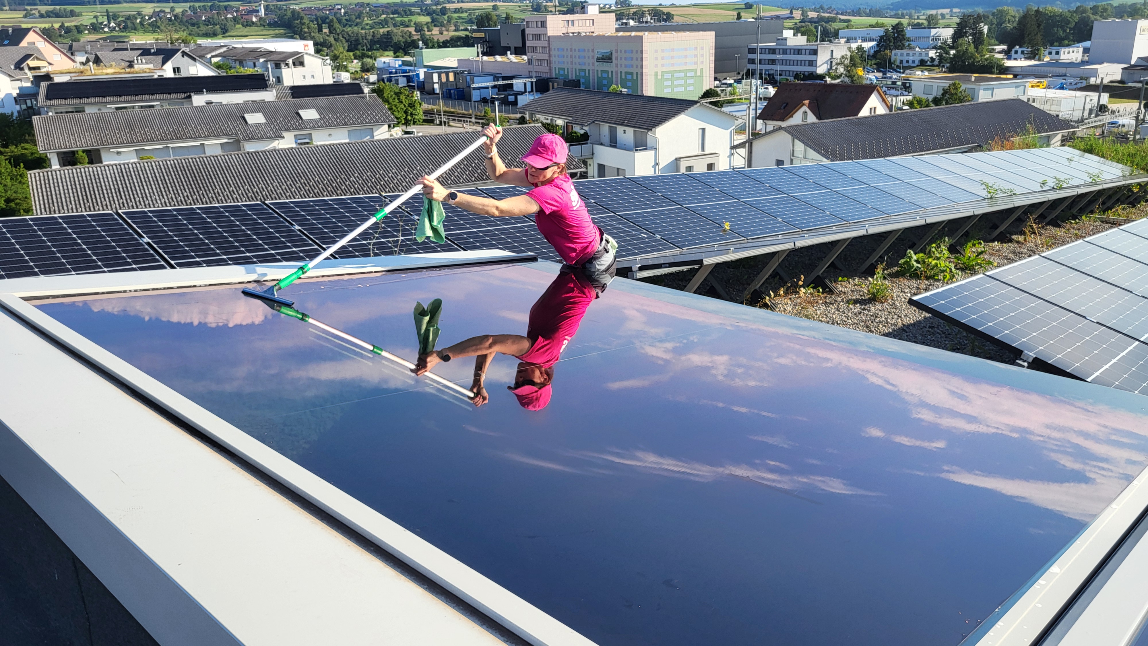 Frau reinigt grosse Solarpanel-Fläche auf einem Dach mit einem langen Wischer, im Hintergrund Häuser und weitere Solarpanels.