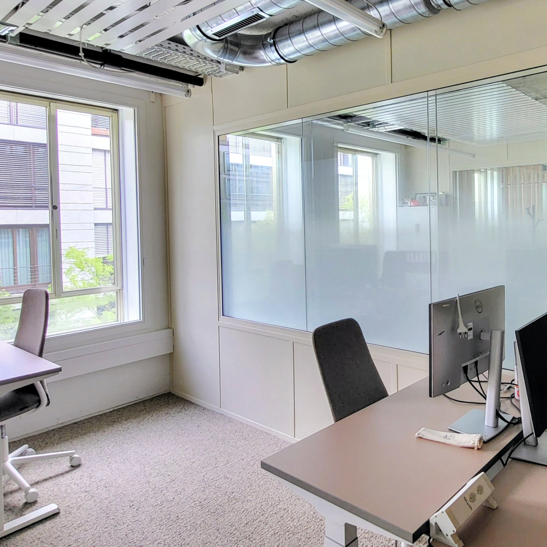 Helles Büro mit zwei Schreibtischen, Stühlen, Monitoren und großen Fenstern mit Blick nach draußen.