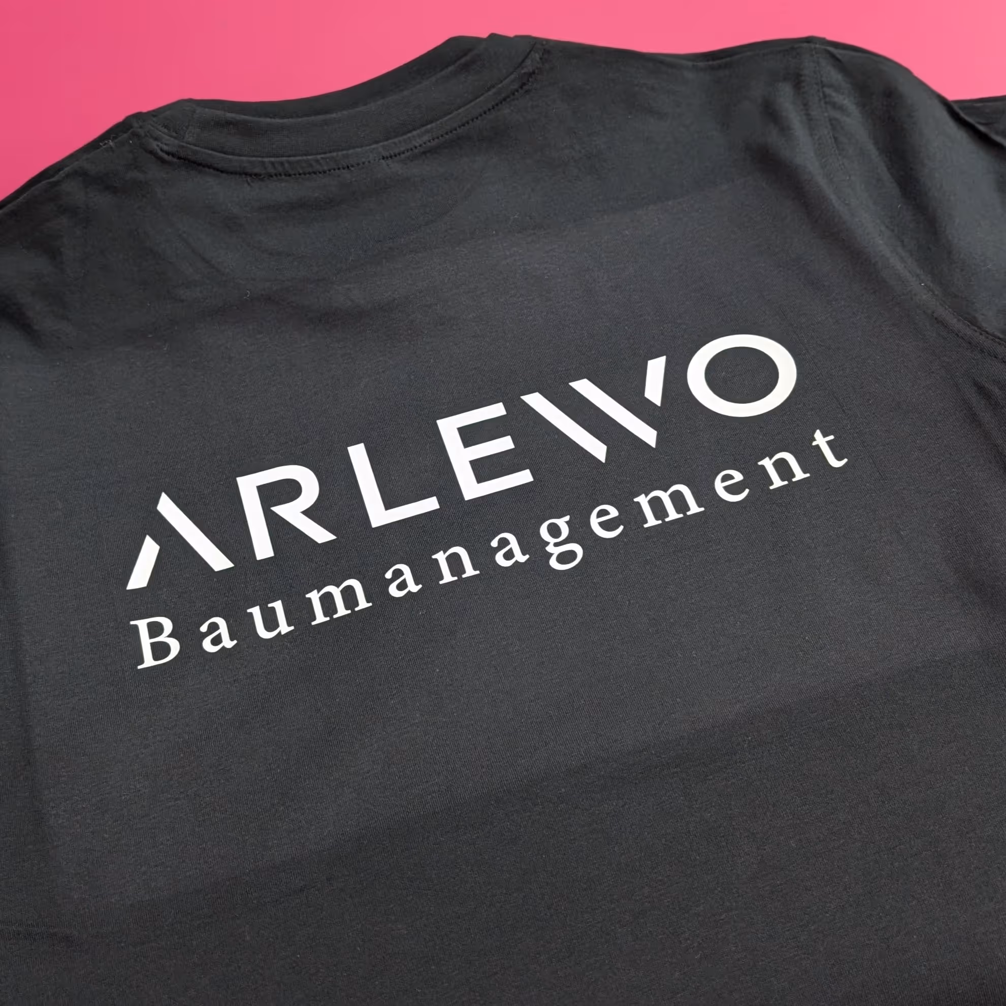 Schwarzes T-Shirt mit weißem ARLEWO Baumangement Aufdruck auf der Rückseite vor rosa Hintergrund.