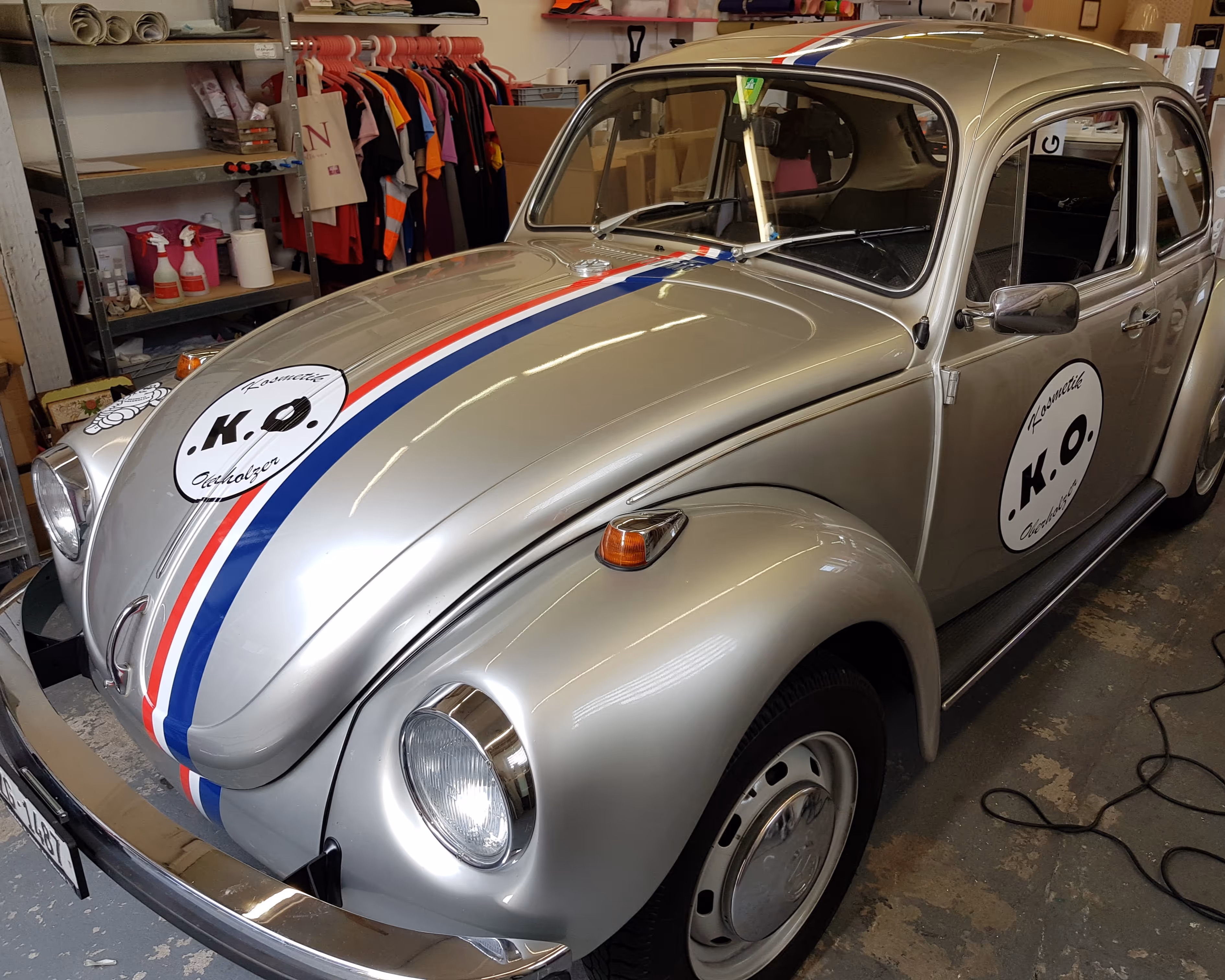 Silberner VW Käfer mit roten und blauen Streifen und K.O. Aufklebern in einer Werkstatt.