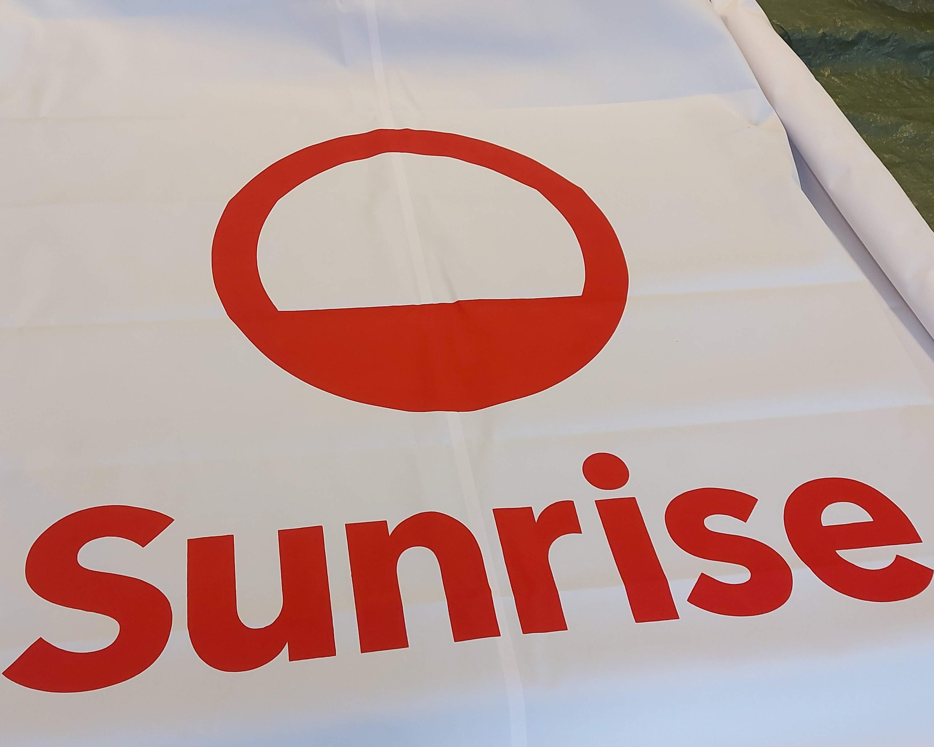 Ein modernes Gebäude mit braunen Fassadenelementen und großen Fenstern; darunter stehen zwei Personen freudig auf einem glänzenden Solarpaneldach unter klarem blauem Himmel.