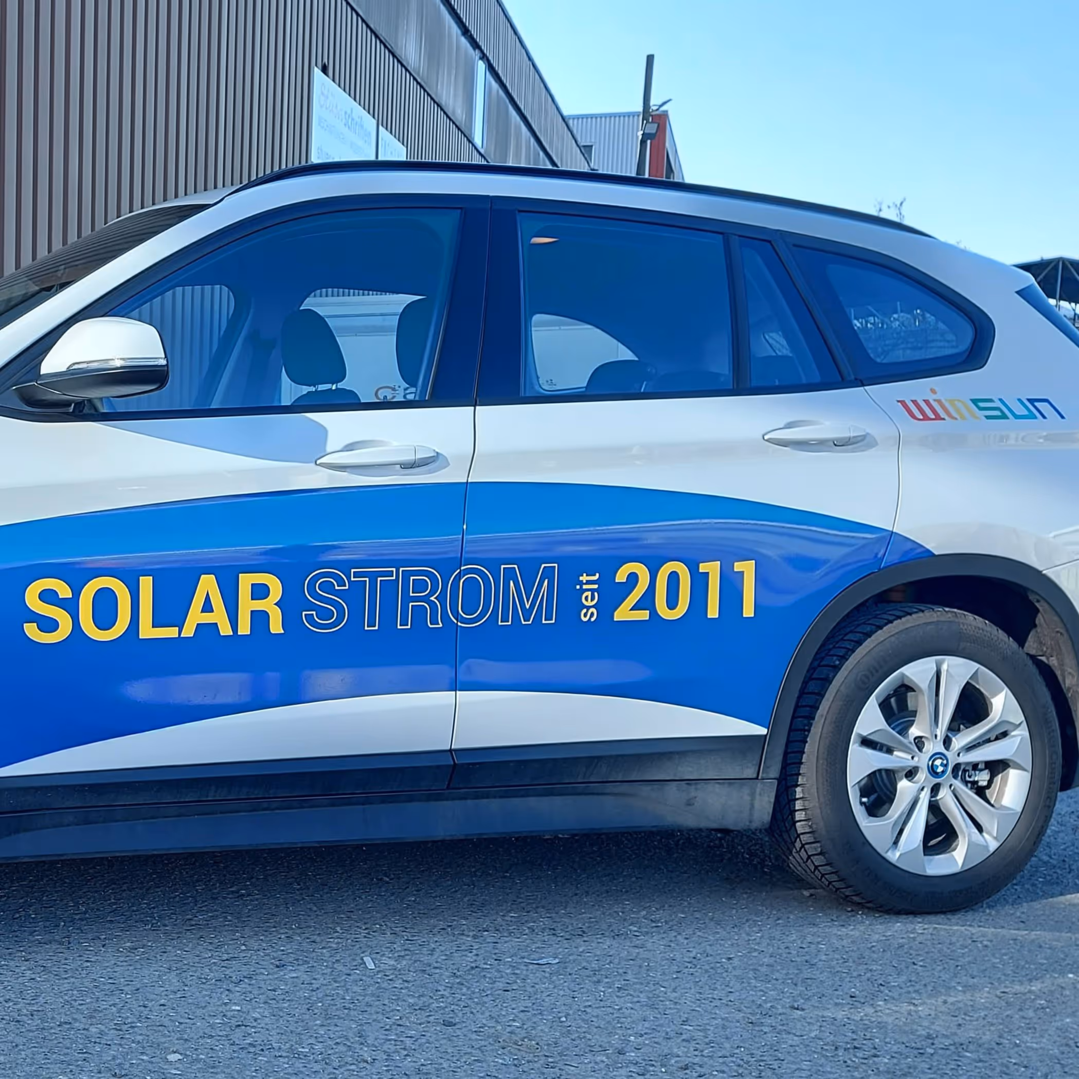 Ein modernes Gebäude mit braunen Fassadenelementen und großen Fenstern; darunter stehen zwei Personen freudig auf einem glänzenden Solarpaneldach unter klarem blauem Himmel.