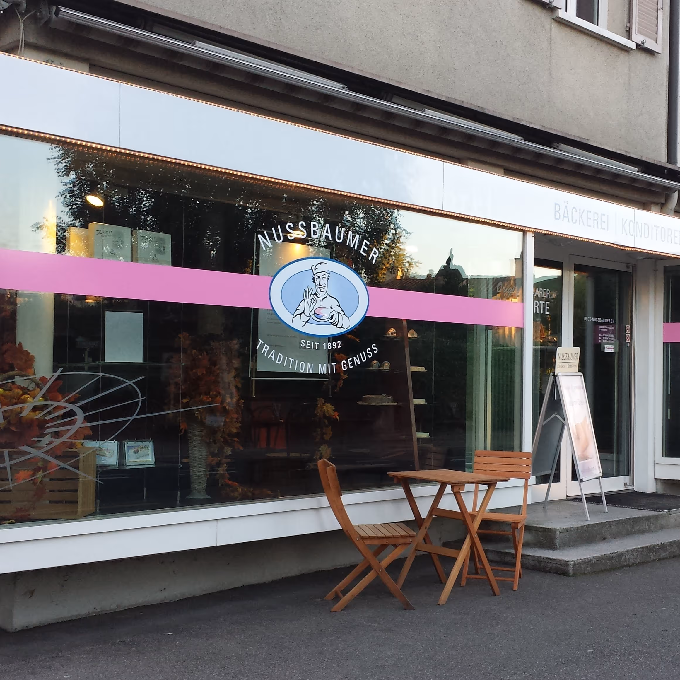 Frontansicht von Nussbaumer Bäckerei mit zwei Holztischen und Stühlen auf dem Gehweg davor.