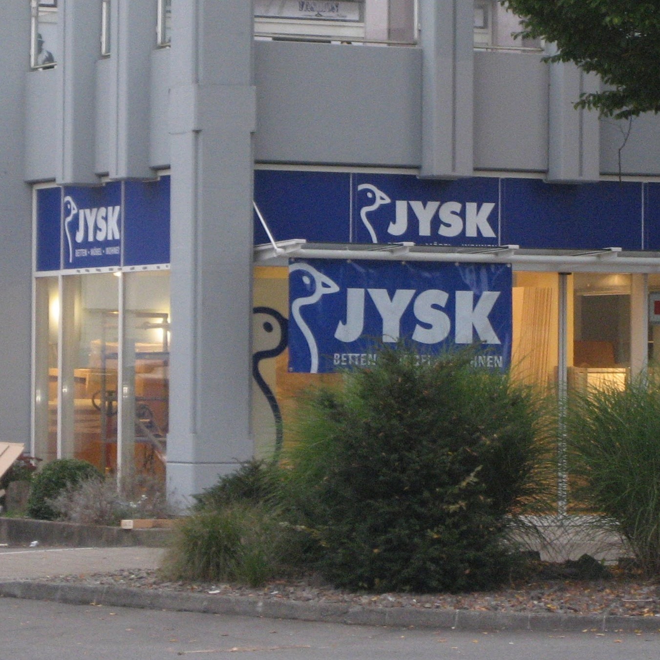 JYSK-Geschäft mit blauen Schildern und Logo, von außen mit Büschen davor.