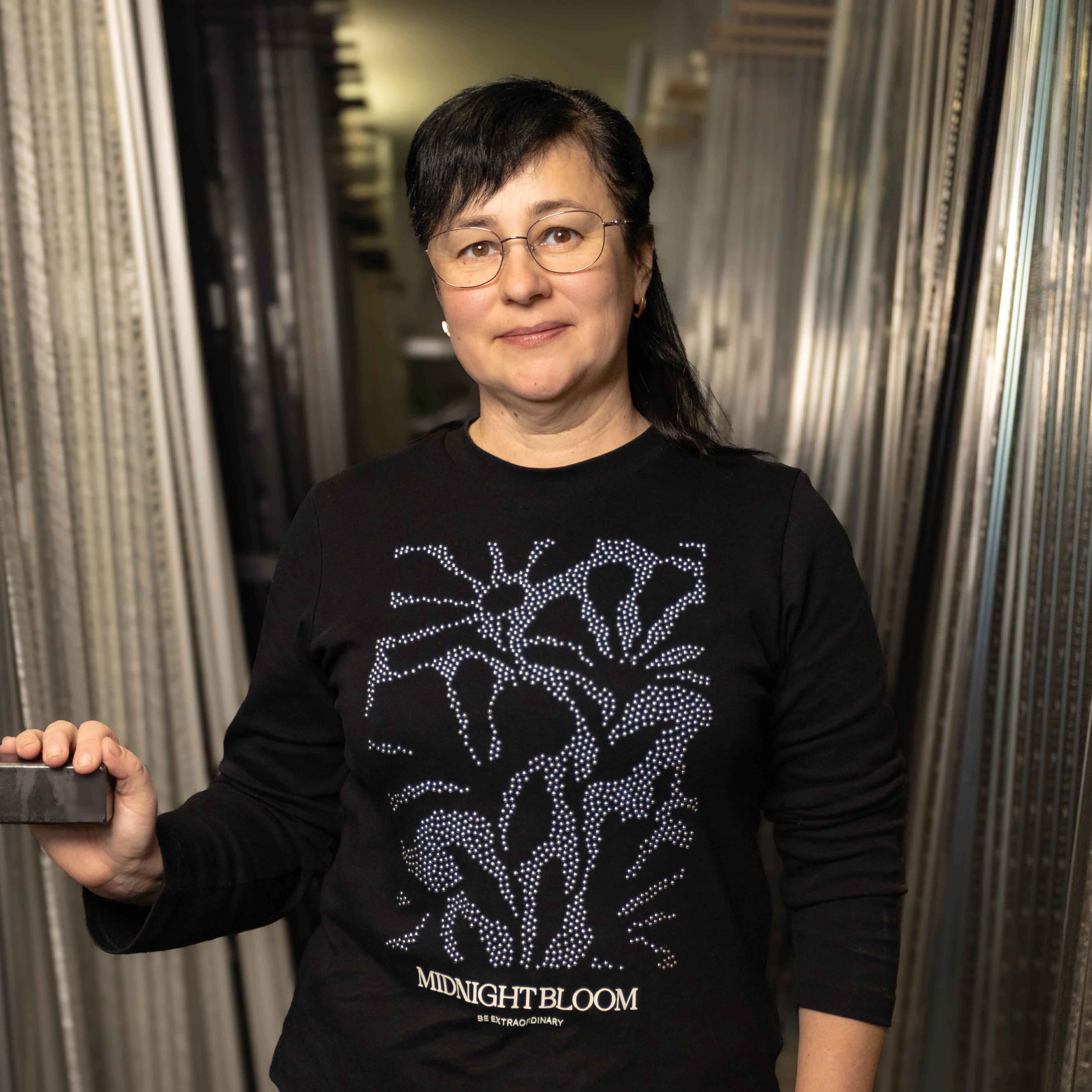 Frau mit schwarzem Pullover mit floralem Muster und Schriftzug „MIDNIGHT BLOOM“, die in einem Gang mit Metallregalen steht.