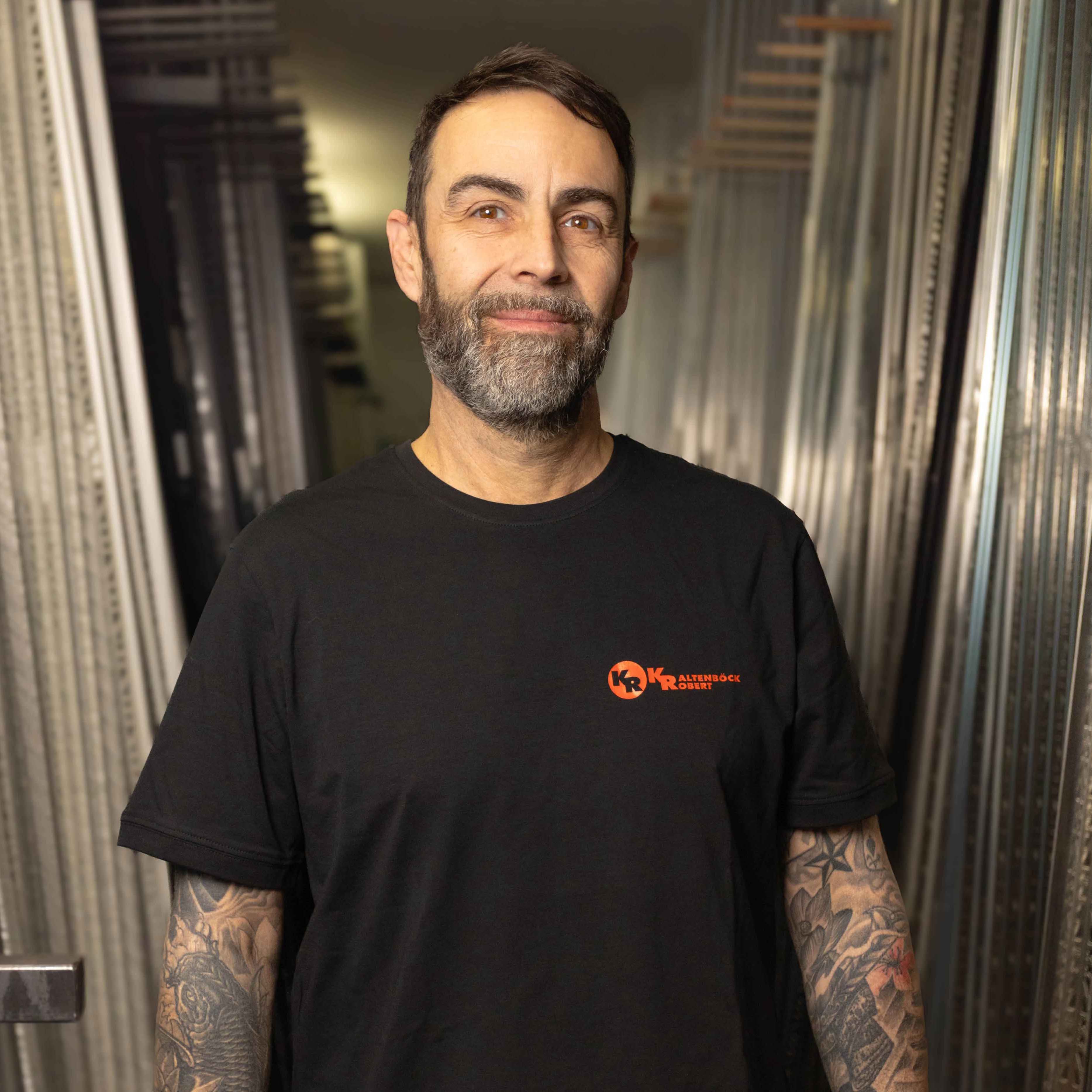 Mann mit Bart und Tattoos an den Armen trägt ein schwarzes T-Shirt mit orangefarbenem Logo in einer industriellen Umgebung.
