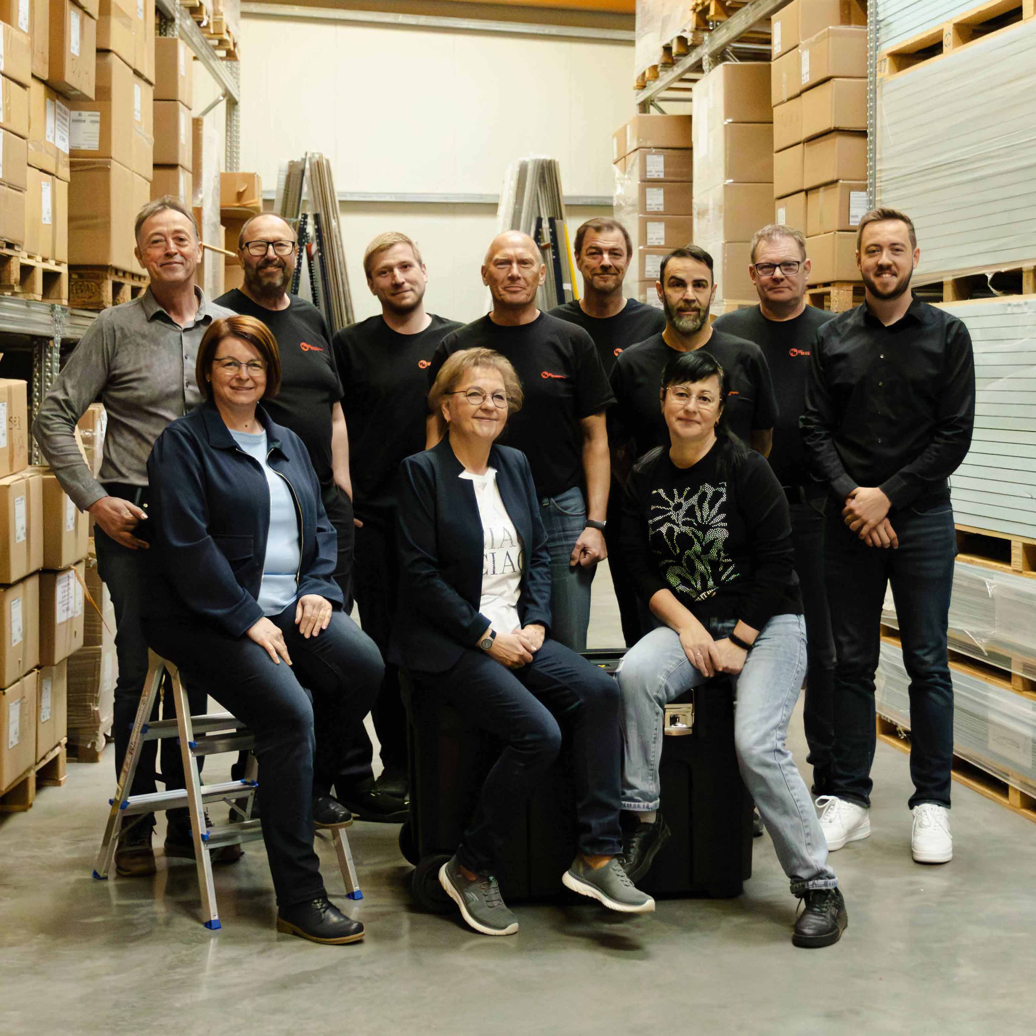 Gruppenfoto von elf Personen in einem Lager mit Kartons auf Regalen, einige sitzen und andere stehen, die meisten tragen schwarze Shirts mit Firmenlogo.