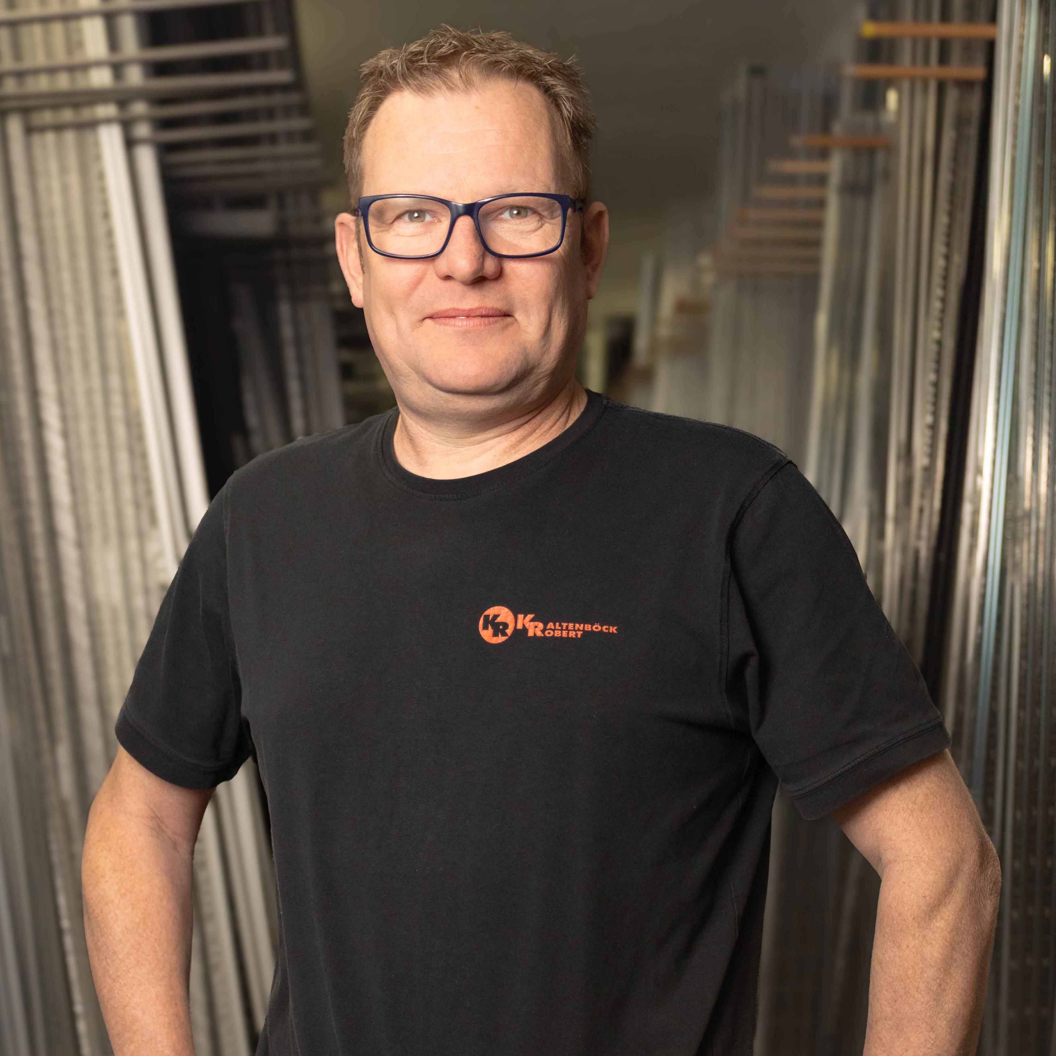 Mann mit blauen Brillen und schwarzem T-Shirt mit orangem Logo steht in einem Lager mit Metallrahmen.