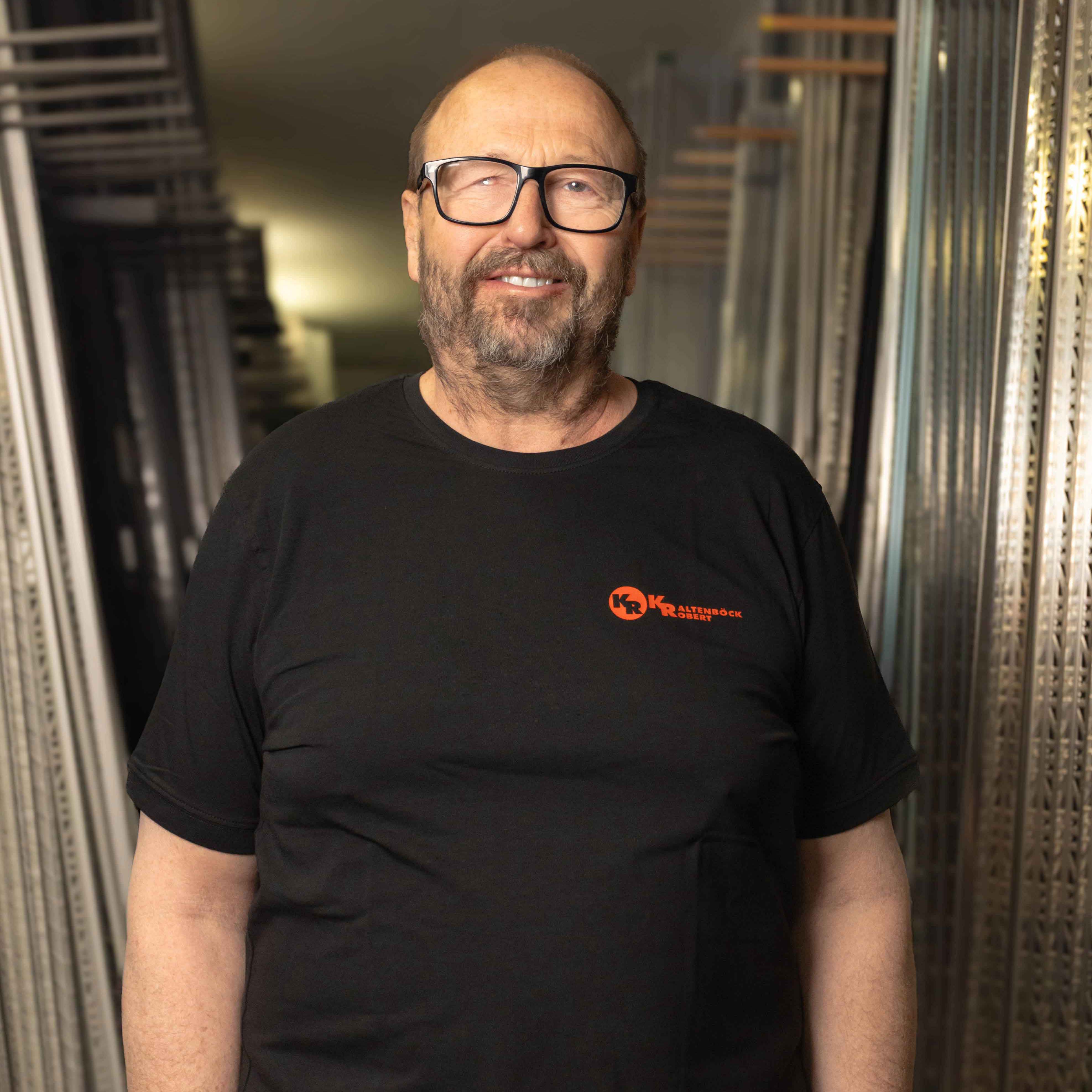 Älterer Mann mit Brille und schwarzem T-Shirt mit orangem Logo steht zwischen Metallregalen in einer Werkstatt.