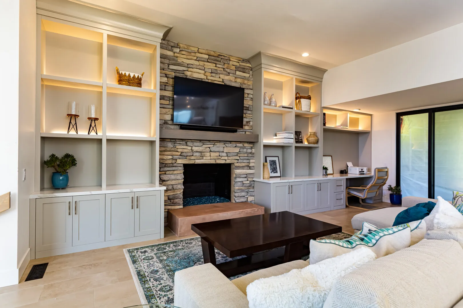 Modern living room with built-in shelving and cabinets, a stone fireplace with a mounted TV, a dark wooden coffee table, and a light sectional sofa with decorative pillows.