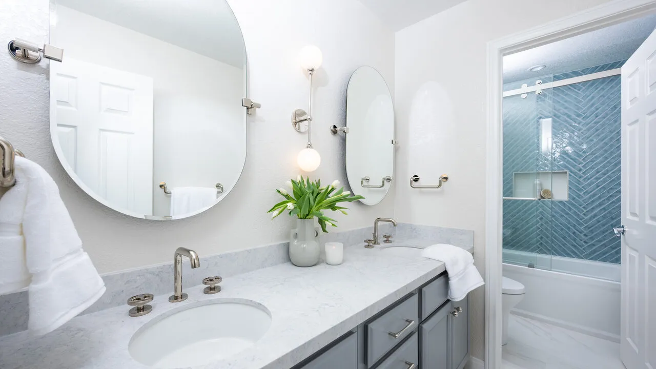 Modern bathroom with double sink vanity, gray cabinets, round mirrors, and a bathtub with blue tiled wall.