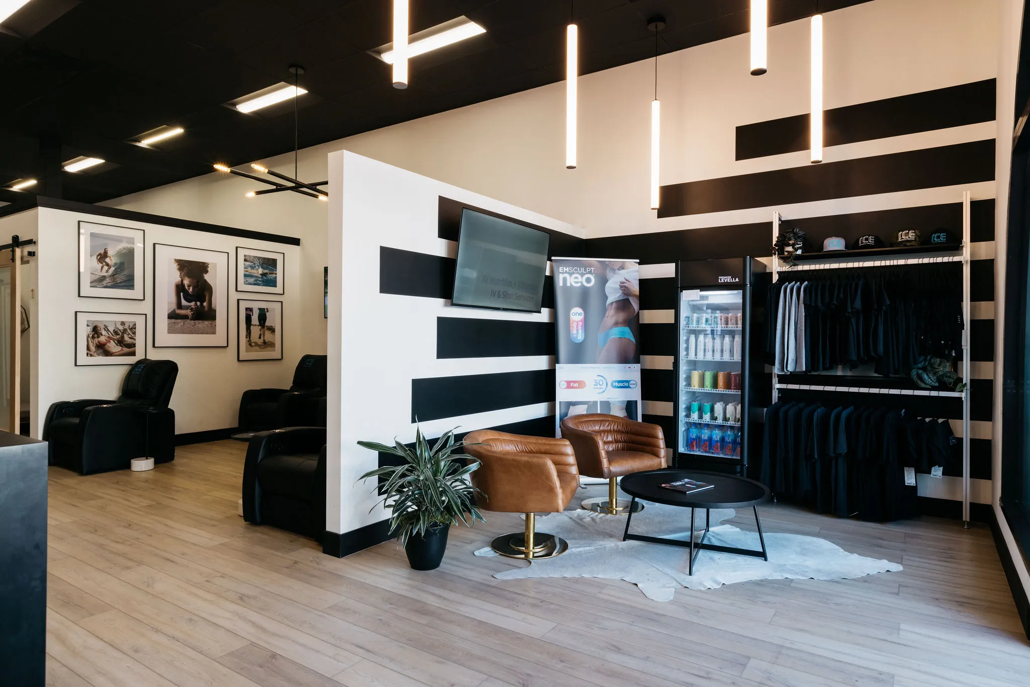 Modern waiting area with two brown leather chairs, a round black coffee table, a potted plant, a refrigerator with drinks, and apparel displayed on shelves.