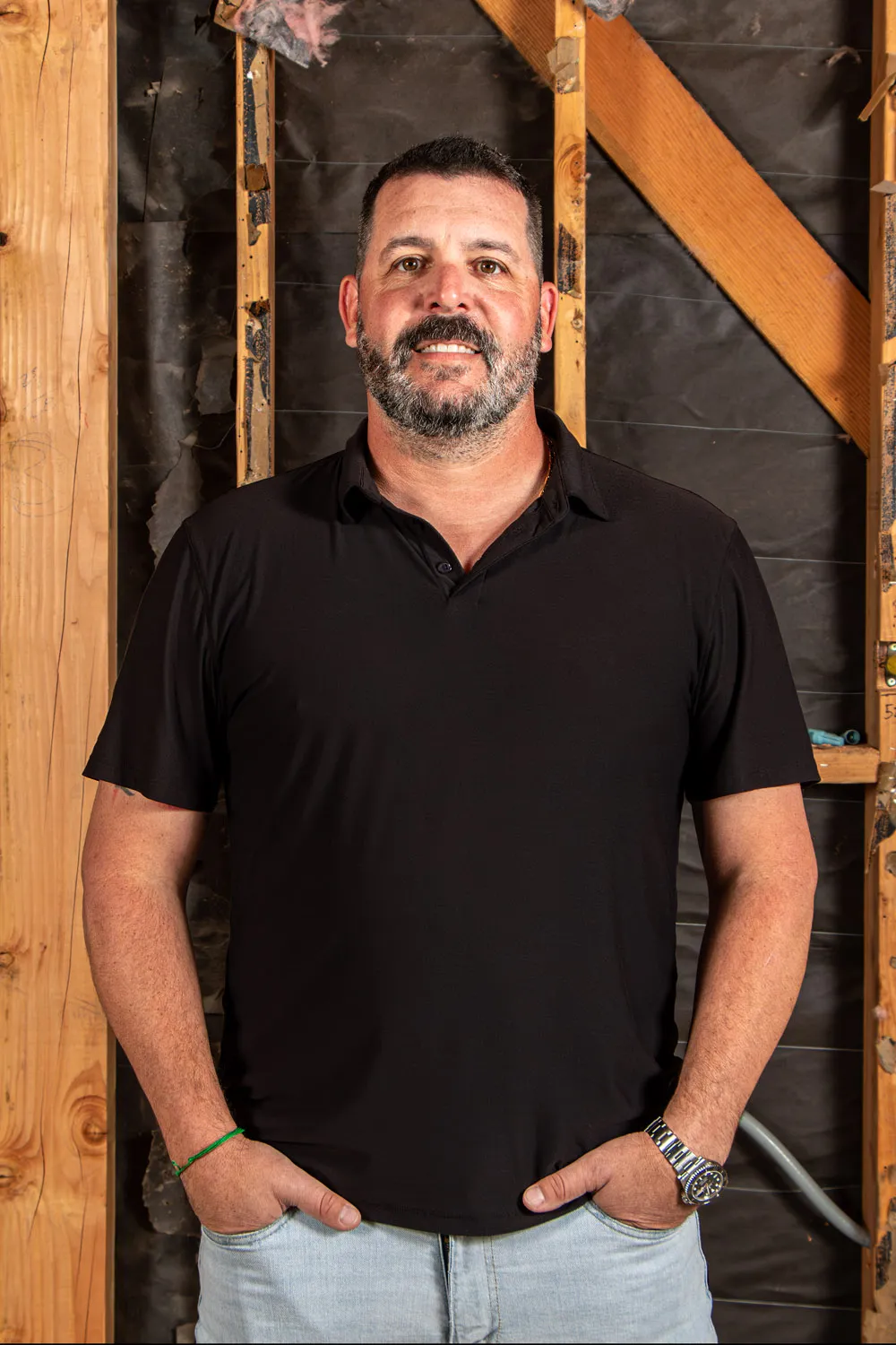 Man with short dark hair and beard wearing a black polo shirt and light jeans standing with hands in pockets against an unfinished wall.