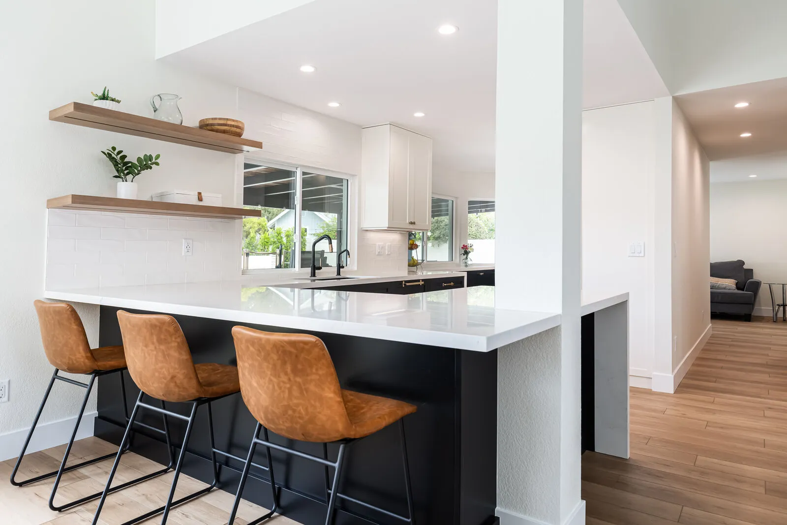 Modern kitchen with a white countertop island, three brown bar stools, open wooden shelves with decor, black faucets, and wood flooring.