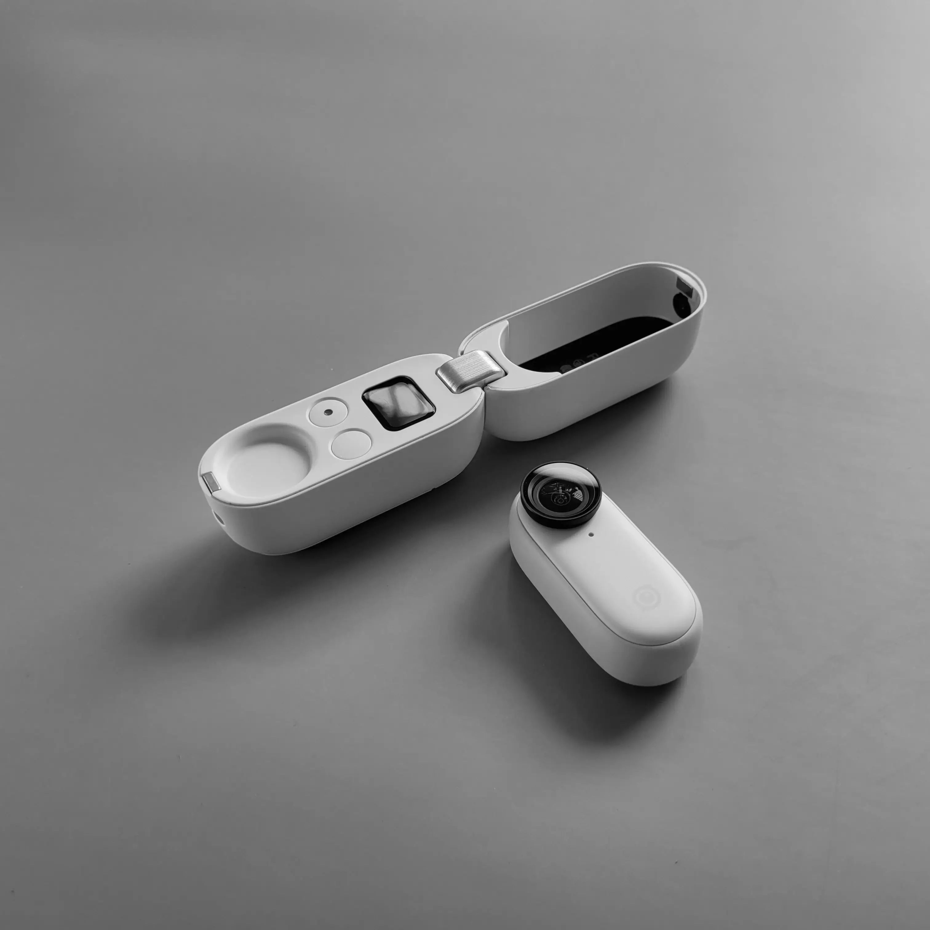 Compact action camera with a detachable lens and an open white charging case on a gray surface.