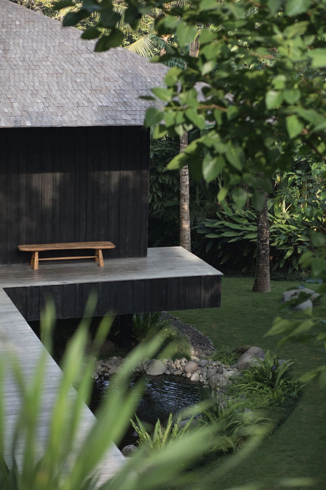 Dark timber exterior with a simple wooden bench on an elevated platform, surrounded by greenery.