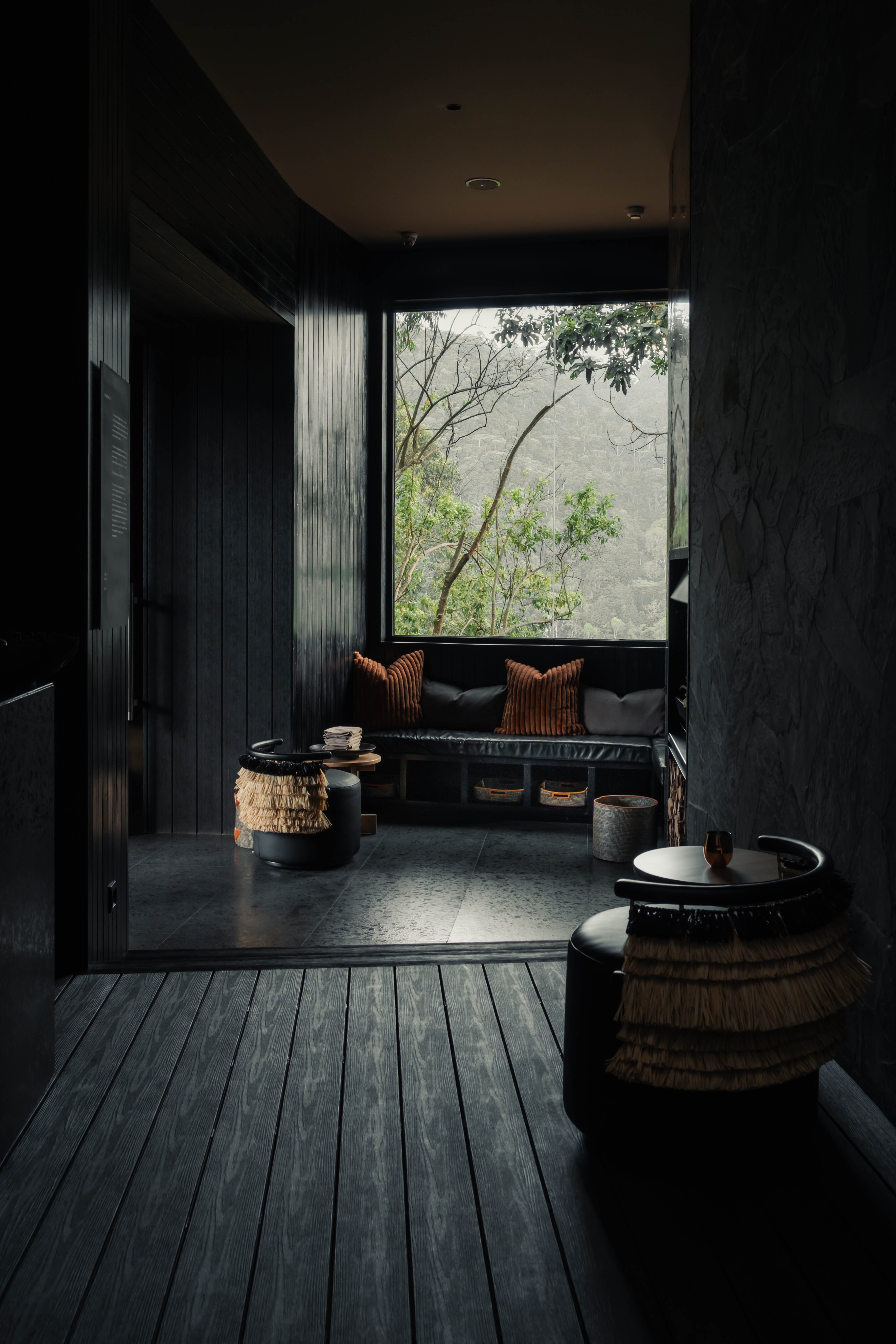Dark modern room with bench seating, brown cushions, small tables, and a large window showing trees outside.