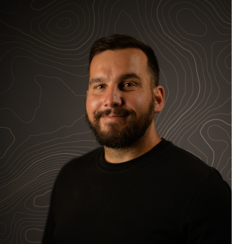 Smiling man with short dark hair and beard wears black shirt against dark topographic map background.