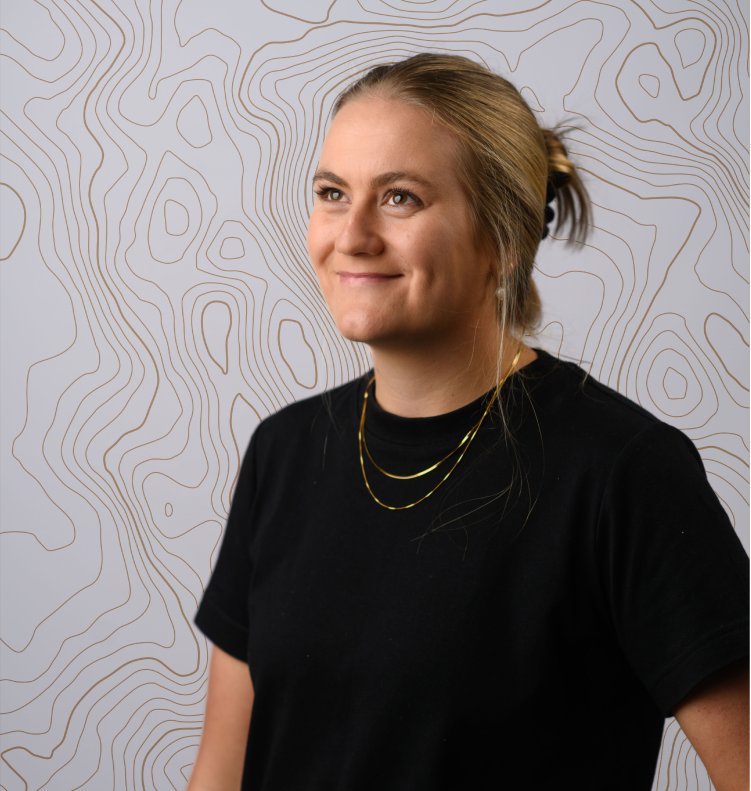 Smiling woman with brown hair tied back, wearing a black shirt and gold necklaces against a line art backdrop.