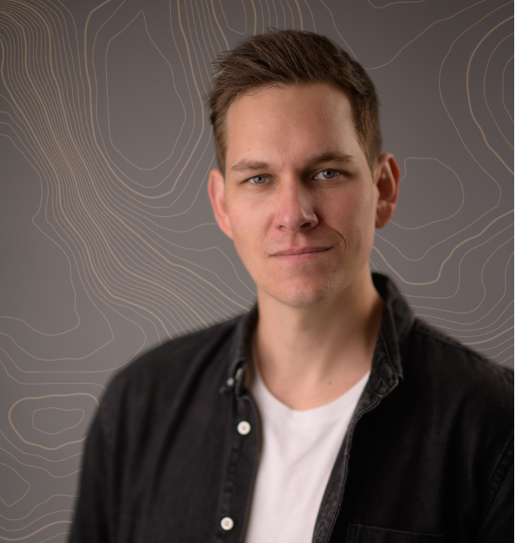 Man with short brown hair wearing a white shirt and black jacket against a gray backdrop with contour lines.