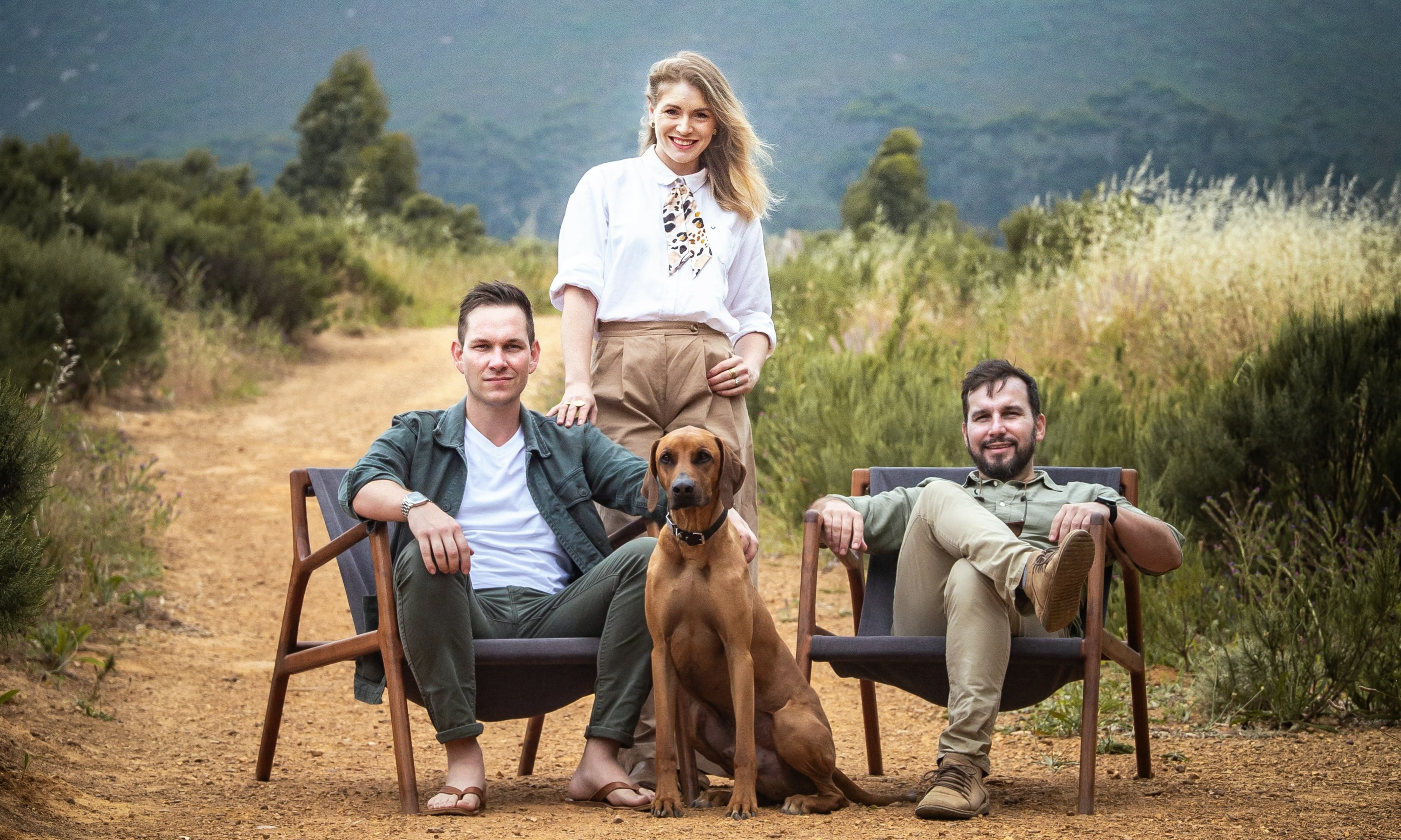 Two men sitting on chairs with a woman standing behind them and a brown dog seated in the middle outdoors.