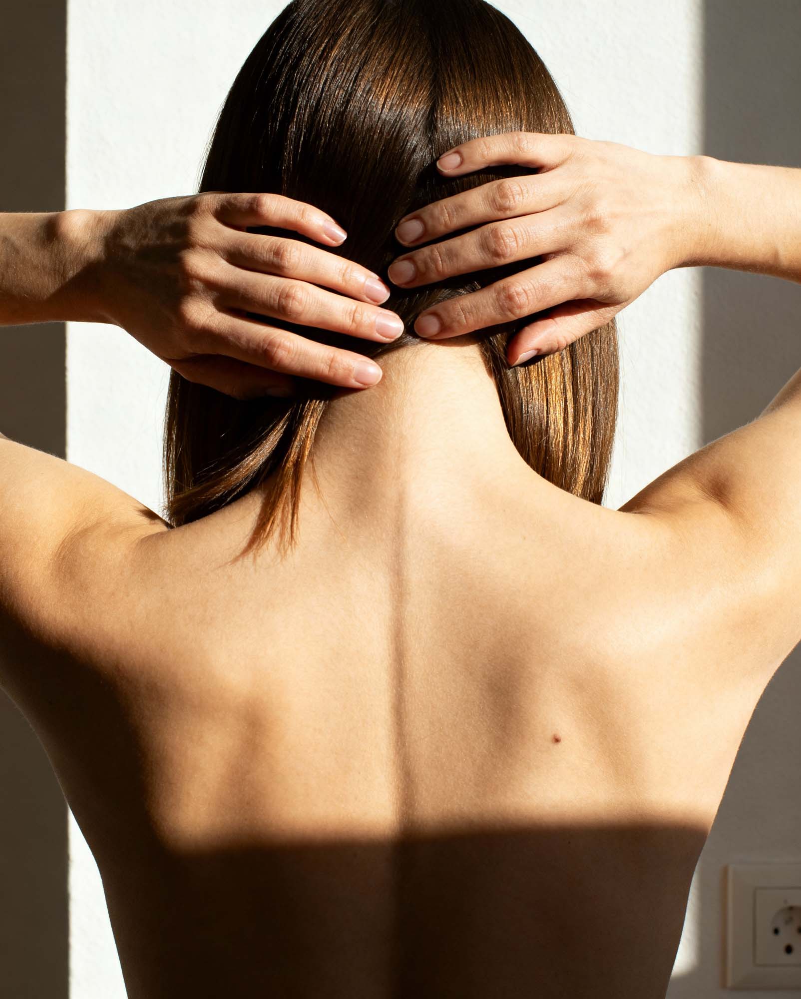 Rücken einer Person mit braunen Haaren, die Hände am Kopf im Sonnenlicht an einer weißen Wand.