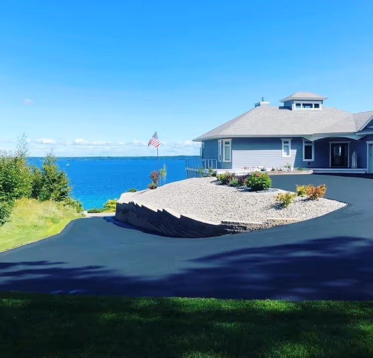 High-end residential asphalt driveway with a fresh, jet-black sealcoat finish and stone retaining wall overlooking the water in Traverse City, Michigan, showcasing Grand Traverse Sealcoating's professional pavement preservation.