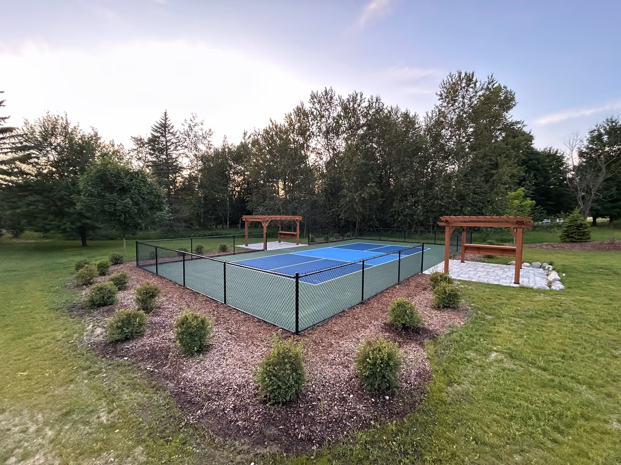 A wide-angle landscape photograph of the completed cushioned pickleball court in Glen Arbor, MI, on Dunns Farm Rd. The court features a high-performance, two-tone blue and green SportMaster surface system with distinct white line striping, fully completed landscaping with mulch and shrubs, and is enclosed by a black chain-link fence between two rustic wooden pergolas. The scene is set under a clear blue sky.