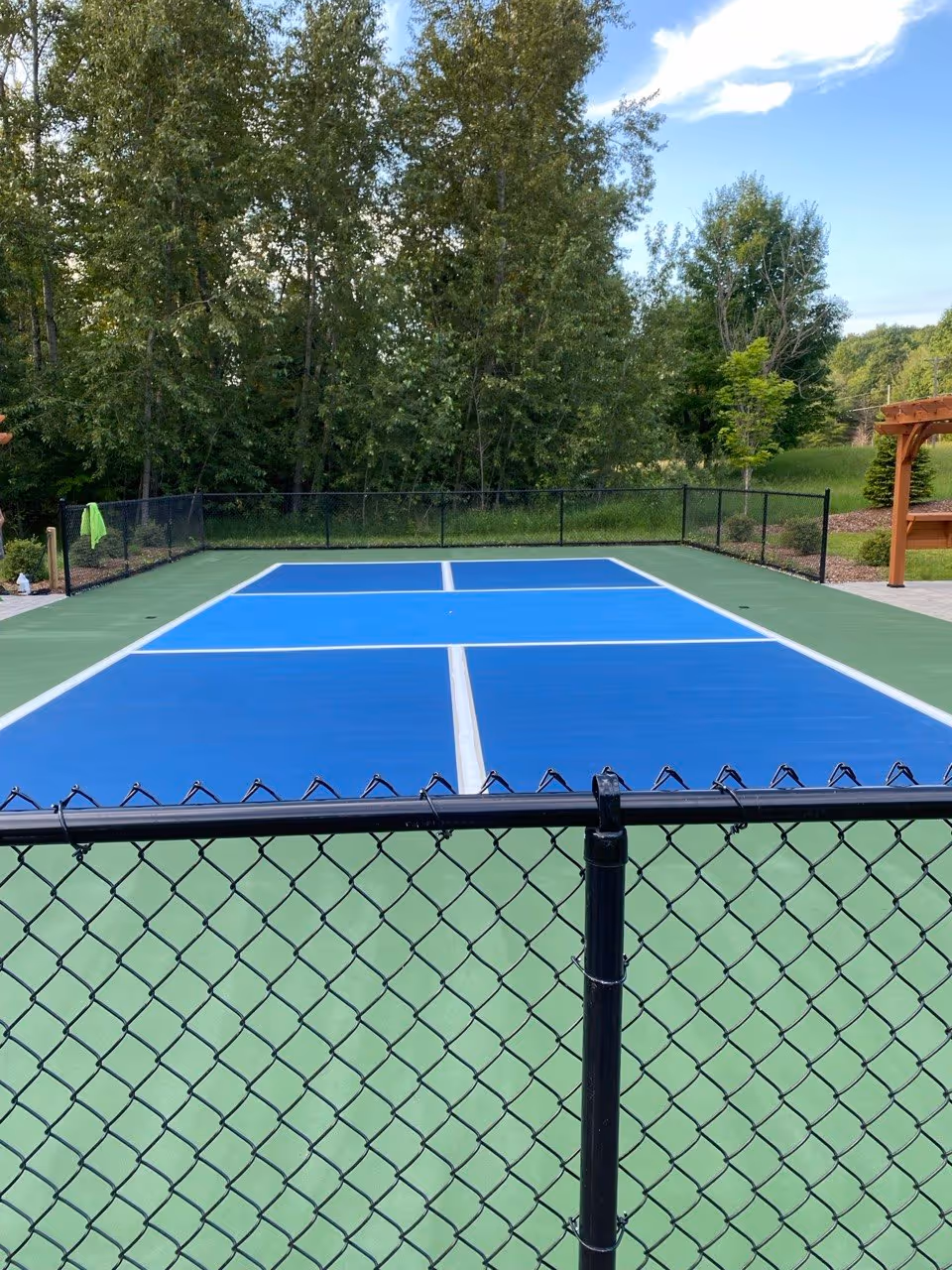 An angled action shot of the precision line striping process. The SportMaster textured acrylic line paint is applied between protective tape lines to create high-visibility white boundary and kitchen lines. Protective plastic sheeting is laid over the newly colored blue sections, and multiple paint buckets are visible along the court edge, ensuring professional tournament-spec accuracy.