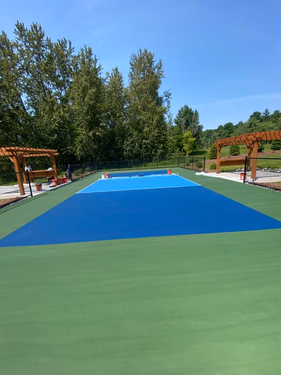 An eye-level photograph from behind the fence, showing the finished playing surface. The private cushioned pickleball court on Glen Lake features a rich navy blue outer boundary, a light blue non-volley zone (kitchen), and clean white lines. The high-grip SportMaster acrylic finish is vibrant and ready for play.