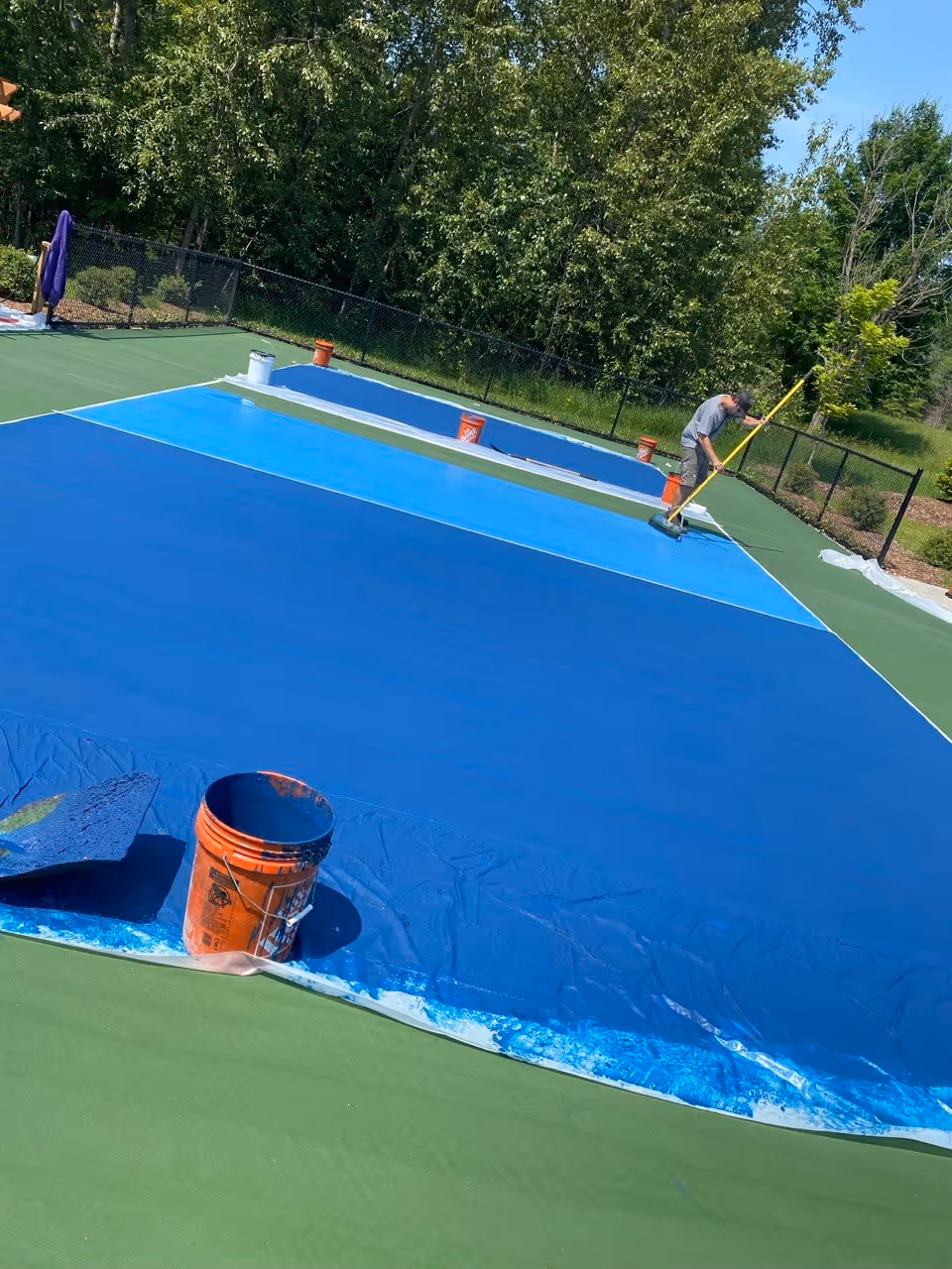 A dynamic action shot showing a crew member in a grey shirt using a specialized squeegee to apply the second vibrant blue SportMaster color coat for the pickleball kitchen area (non-volley zone). The darker navy blue outer boundaries are already complete, and the green baseline area is visible. The sequence highlights the intricate multi-color cushioned surfacing process.