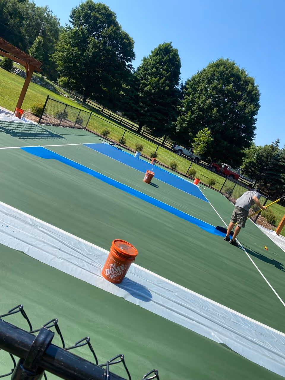 An action photograph showing the application of the primary vibrant SportMaster acrylic color coating. A crew member uses a professional squeegee to achieve a smooth, consistent finish across the cushioned base. Multiple paint buckets containing the specialized joint-friendly surfacing material are positioned along the boundary.