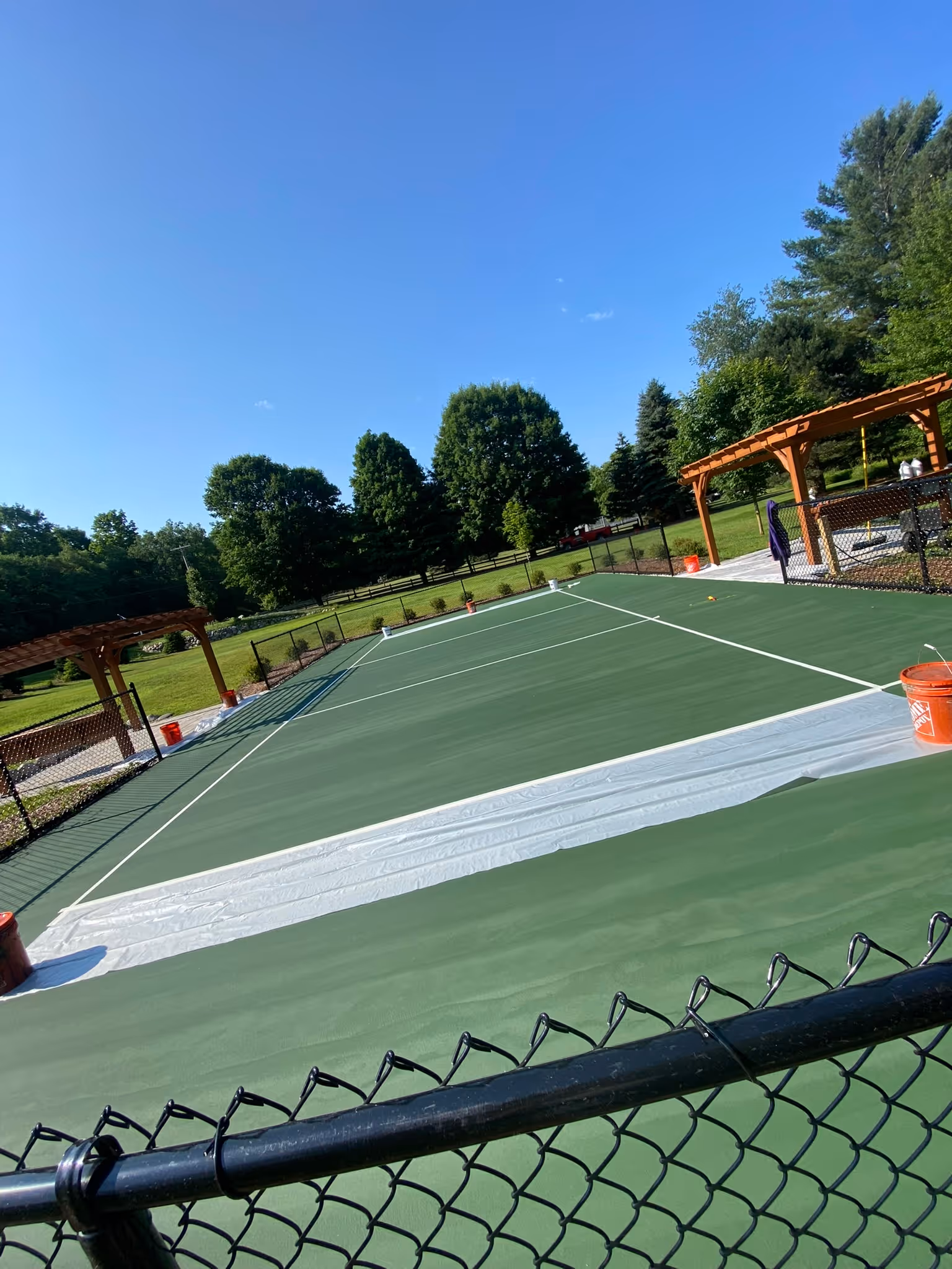 An angled photograph of a crew member in a grey shirt applying the first application of vibrant green SportMaster color coating to the perimeter of the private cushioned court. The grey acrylic resurfacer base is visible where the final color hasn't yet been applied, demonstrating the multi-layer, high-performance cushioned surfacing system.