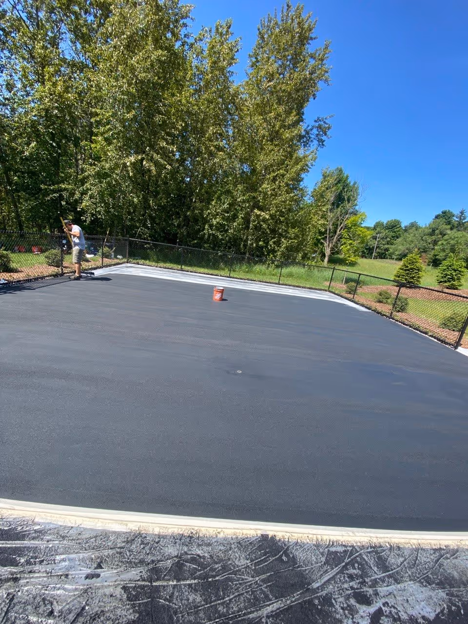 This is a crucial technical photo showing the squeegee application of the specialized SportMaster cushioned rubberized system. A crew member is seen applying the dark rubberized compound over the gray foundation, transforming the raw surface into a joint-friendly, impact-absorbing base. Multiple buckets of surfacing material and a single central red bucket confirm the extensive engineering behind the custom installation on Dunns Farm Rd.