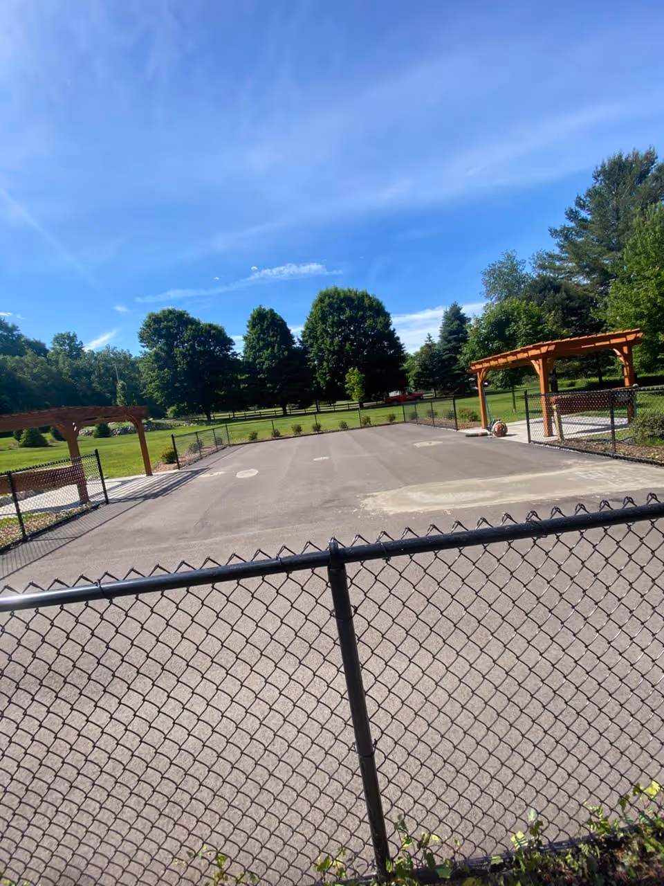The ultimate "before" photo, looking at the raw asphalt court footprint between the pergolas and behind the existing chain-link fence. The scene shows the blank slate before any work began on Dunns Farm Rd. Crucially, a distinct light-grey patch near the foreground confirms that low spot leveling has already been completed, preparing the sub-base for the high-performance cushioned SportMaster surfacing system
