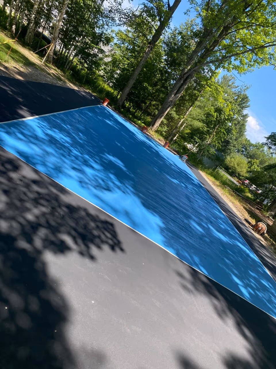 A high-angle view showing the first application of vibrant blue SportMaster acrylic color coating to the internal playing area of a private pickleball court on Dunns Farm Rd. The photo highlights the wet, textured surfacing material being spread across the prepared asphalt base, contrasting against the grey resurfacer near the Big Glen Lake shoreline.